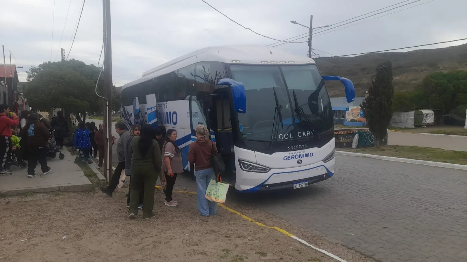 A group of students gathers around a white bus labeled "Transporte Escolar" parked in a school parking lot on a cloudy day.