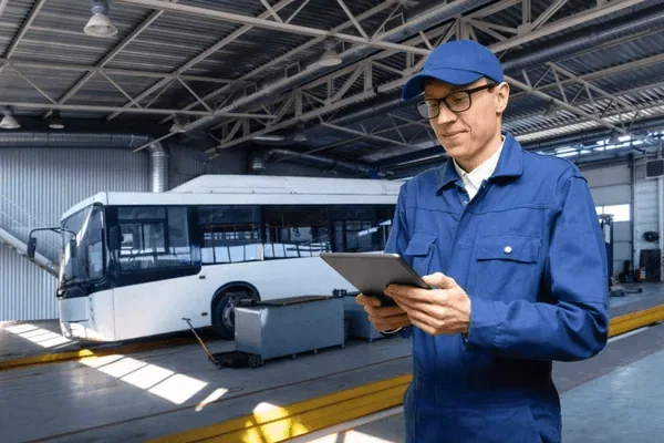 A mechanic in blue overalls and a cap stands inside a spacious, well-lit bus garage, using a tablet to check maintenance records next to a large, white city bus.