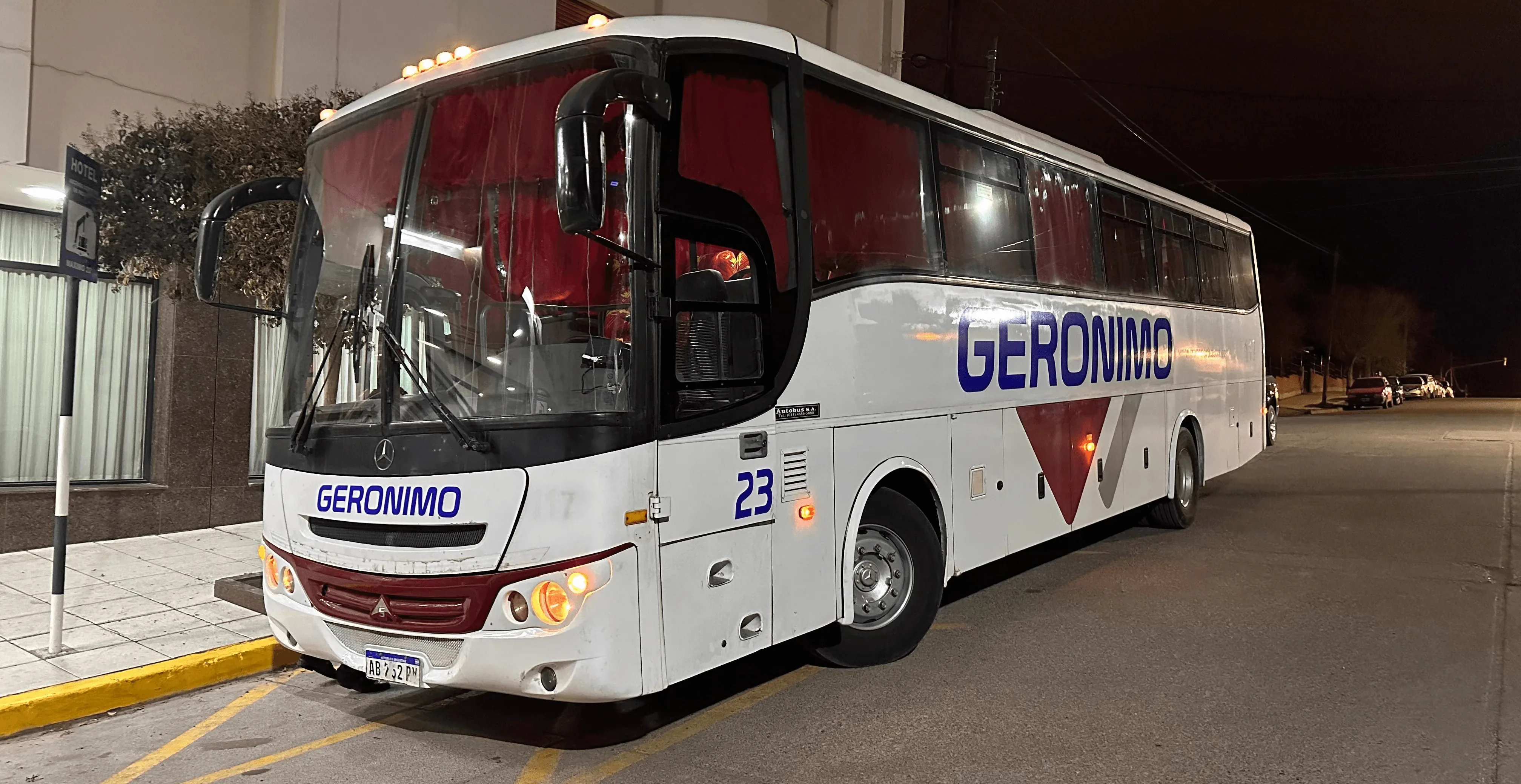 A large white passenger bus with the name "Geronimo" and number "23" on its sides is parked on a well-lit street outside a modern building at night.
