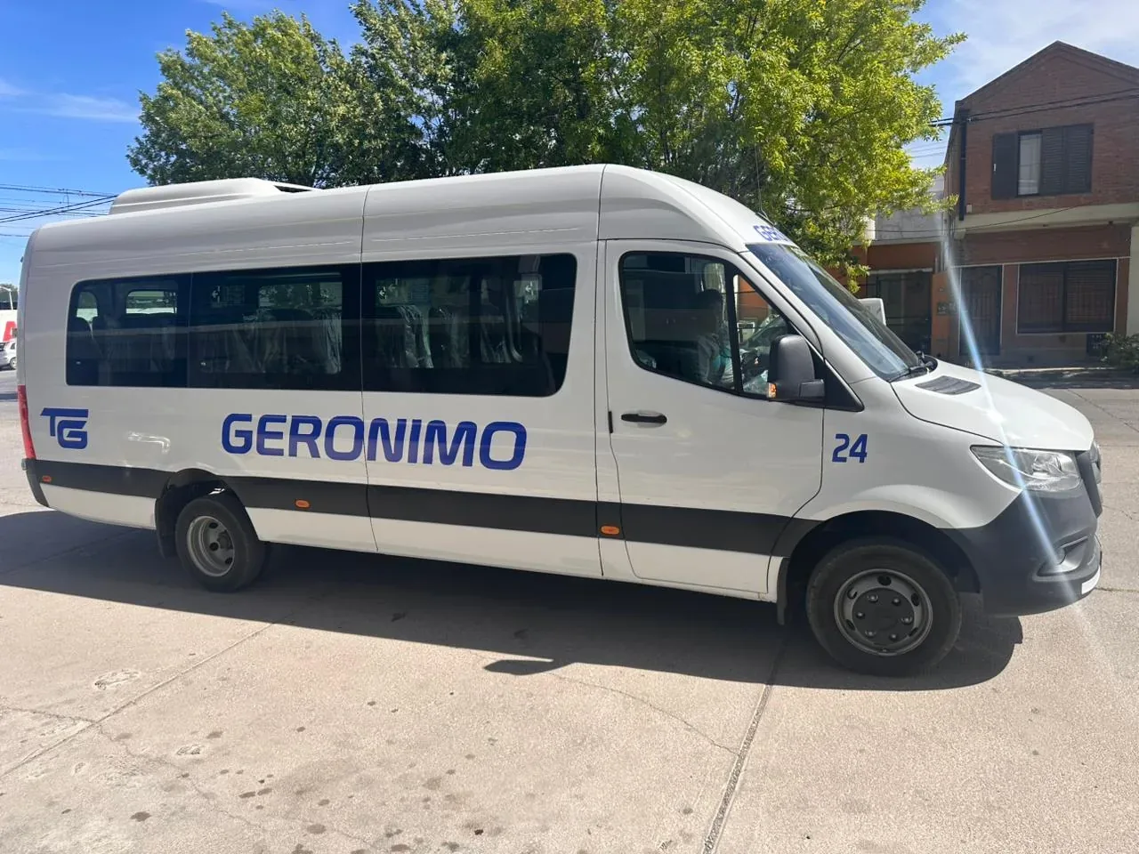 A white passenger van with "Geronimo" written on the side, parked on a sunny street near brick buildings and trees.