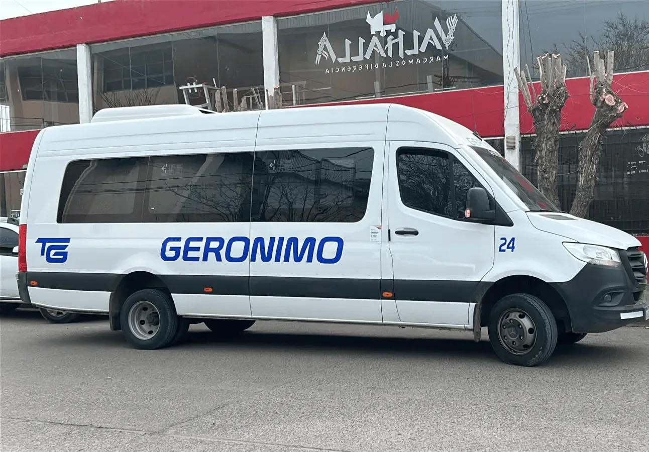 A white transit bus, labeled "Transportes Geronimo," is parked on a dirt road under a partly cloudy sky.