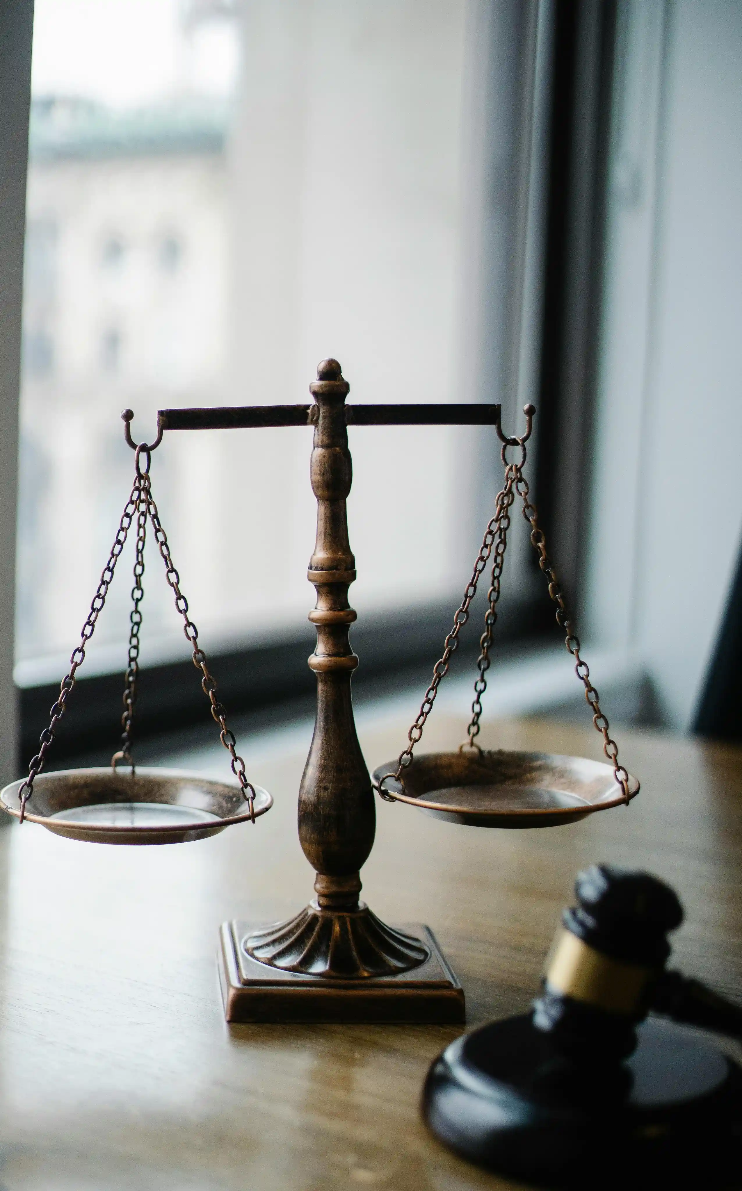 A vintage brass balance scale is placed on a wooden desk next to a judge's gavel, set against a backdrop of a large window revealing an urban cityscape.