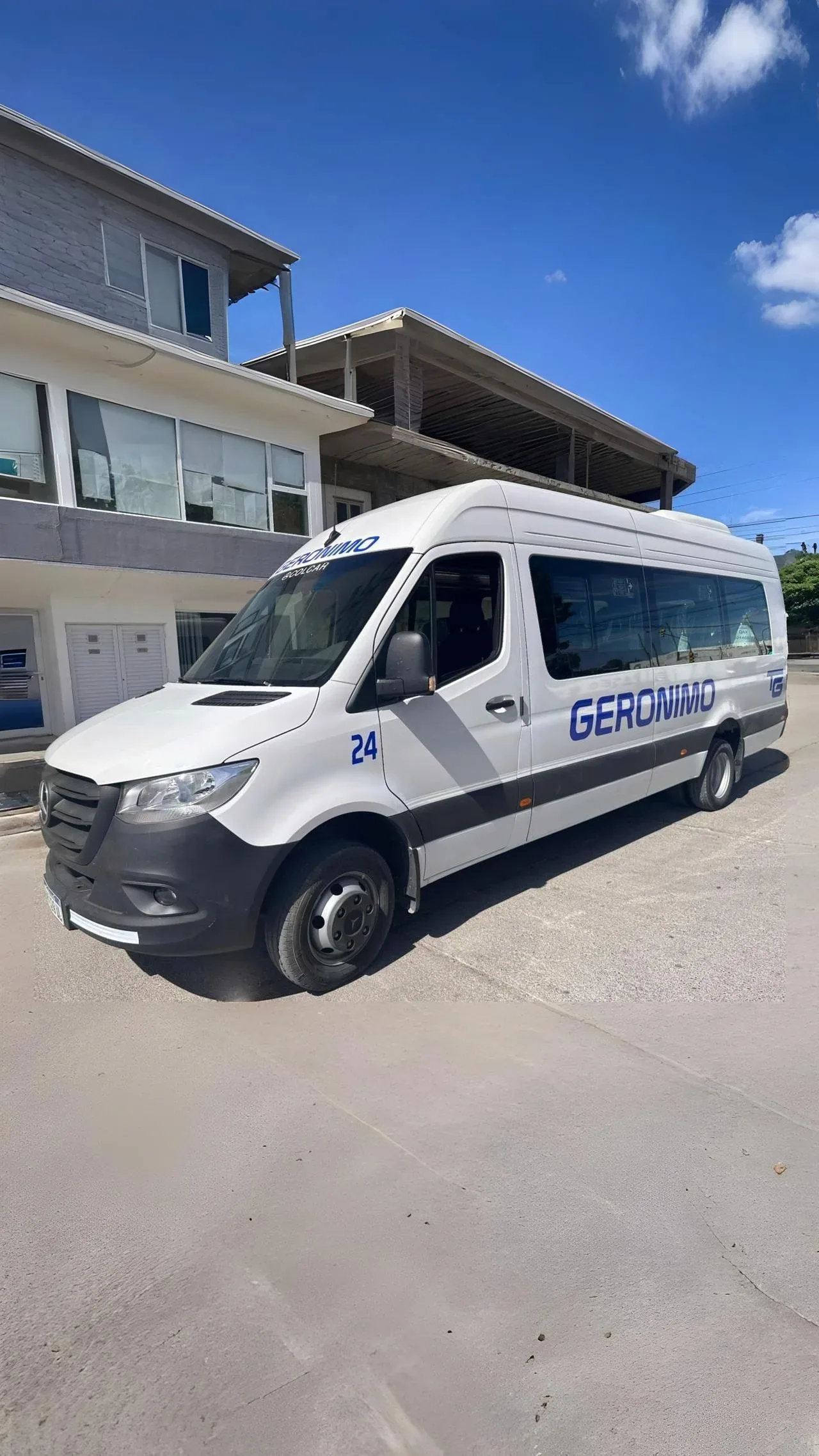 A white passenger van with the text 'Geronimo' on its side is parked on a street in front of a modern building, ideal for group transportation services and featuring sleek design and tinted windows.