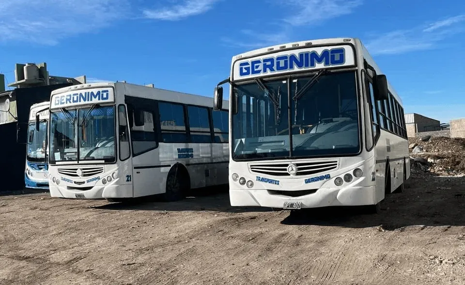 Two white and blue buses with "GERONIMO" signage on the windshield are parked on a dirt lot under a clear blue sky.