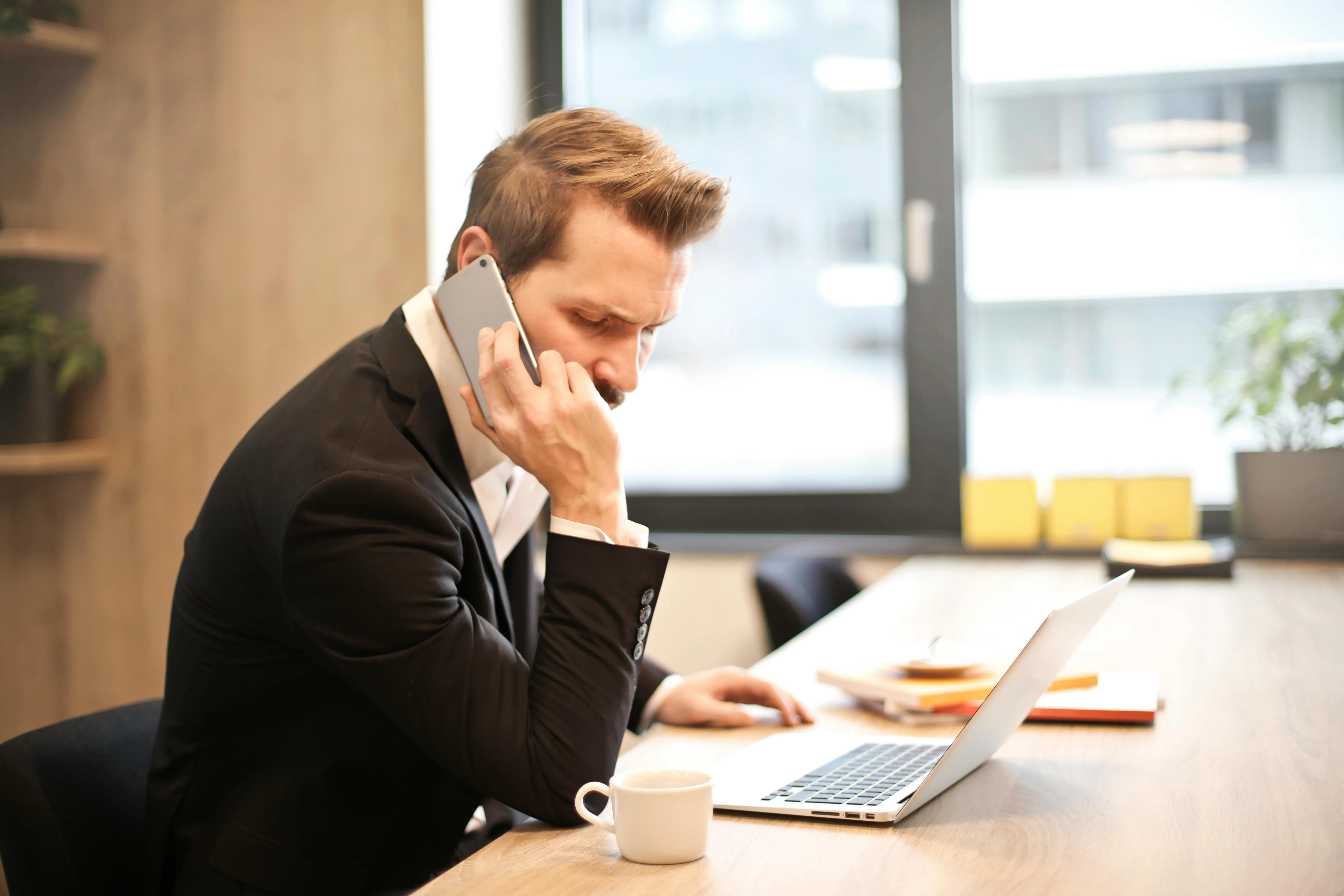 A man in a suit is sitting at a wooden desk, speaking on a smartphone, with a laptop open in front of him, a cup of coffee, and notebooks nearby, in a modern office setting with a large window.