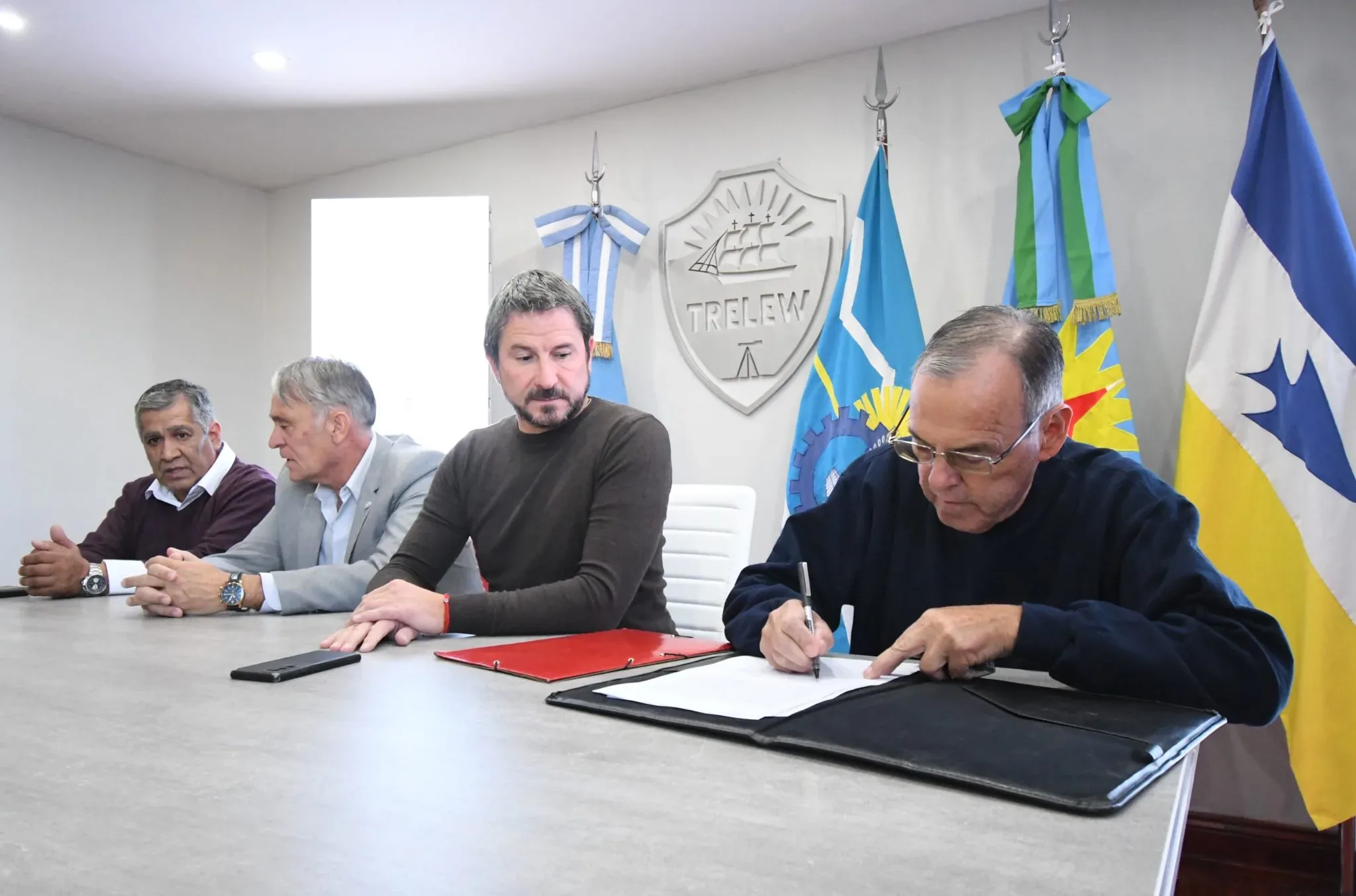 A group of four men are seated at a conference table with one signing a document, while flags and a "TRELEW" emblem are displayed in the background, suggesting an official meeting or agreement signing.