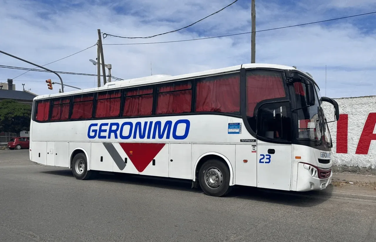 A large white bus with "GERONIMO" written in bold blue letters, featuring maroon accents and tinted red windows, is parked on a paved street, surrounded by urban elements and utility poles under a partly cloudy sky.