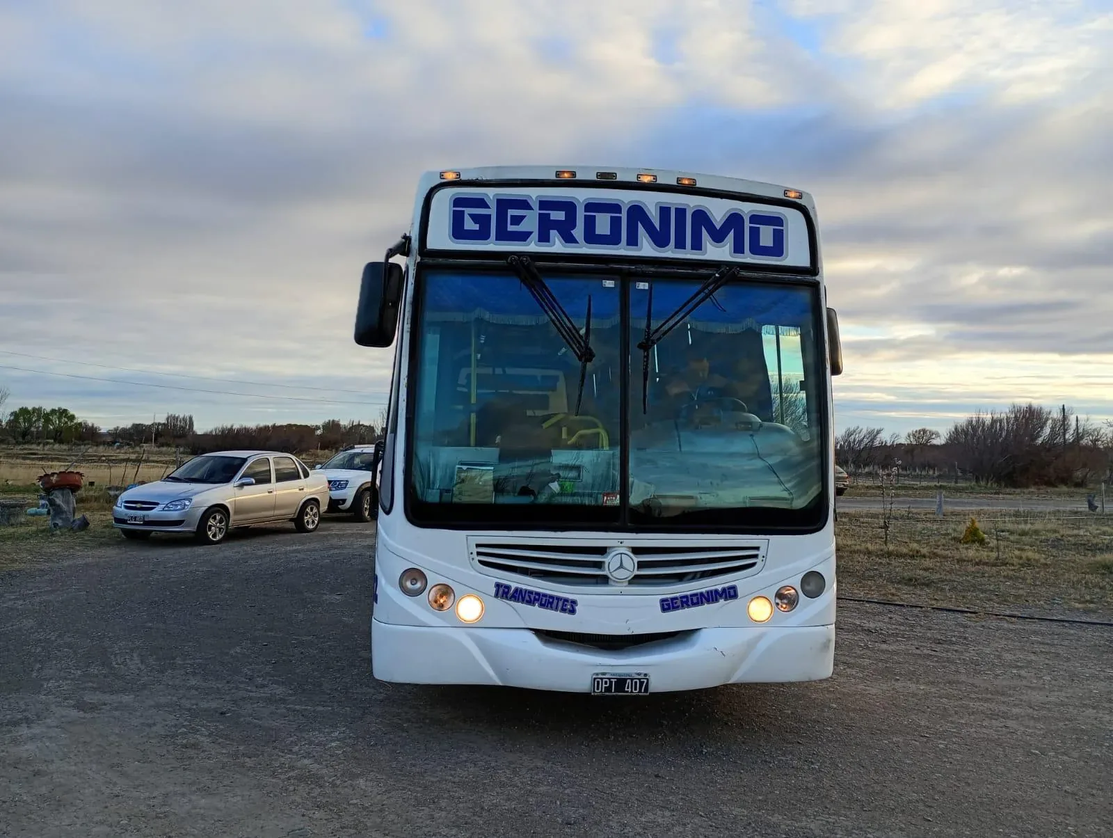 A large white tour bus labeled "Geronimo" is parked on a dirt road, with a cloudy sky and some trees and a car in the background.