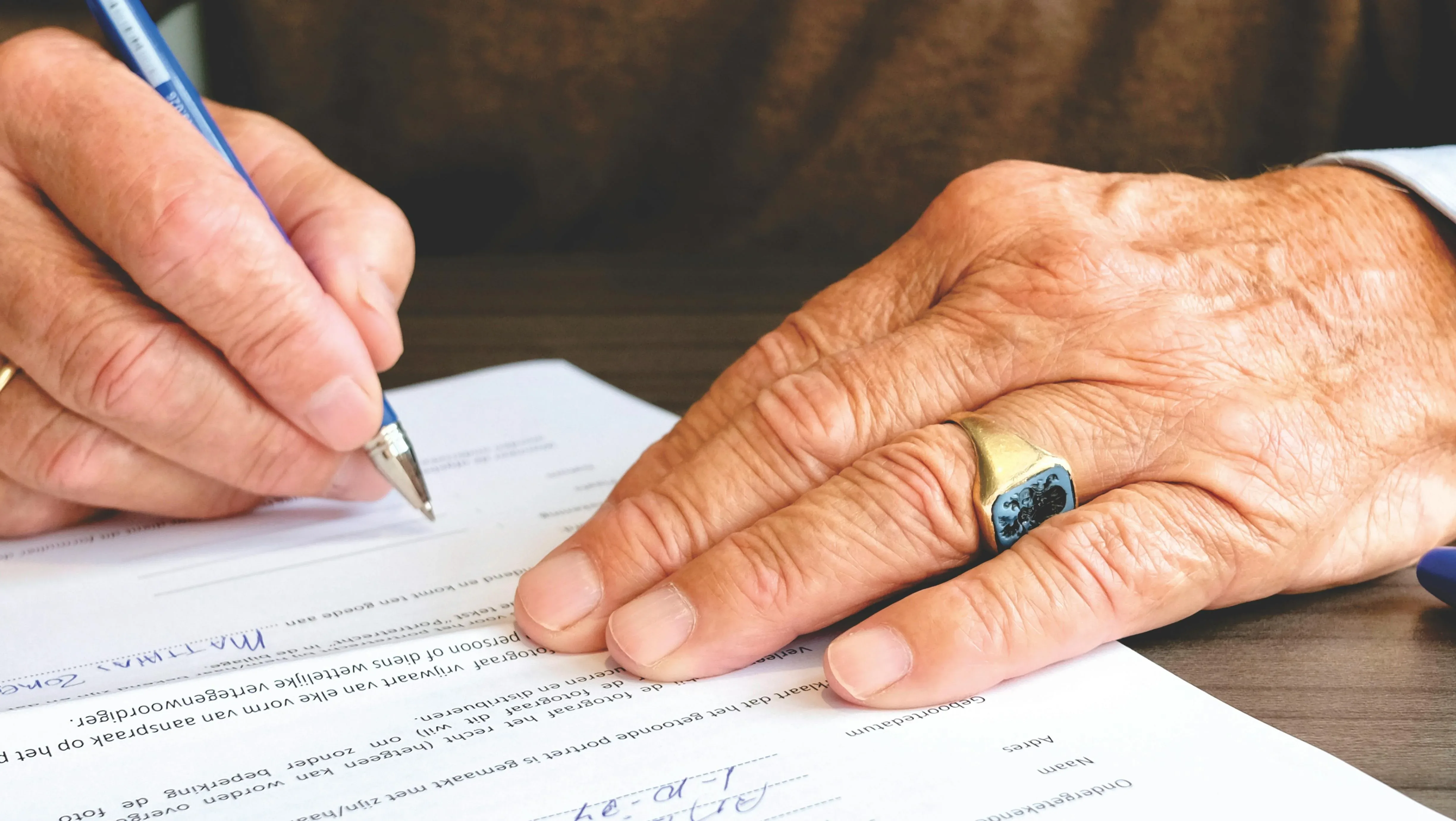 A person wearing a ring is signing a document on a wooden table, using a blue pen, with only their hands visible.