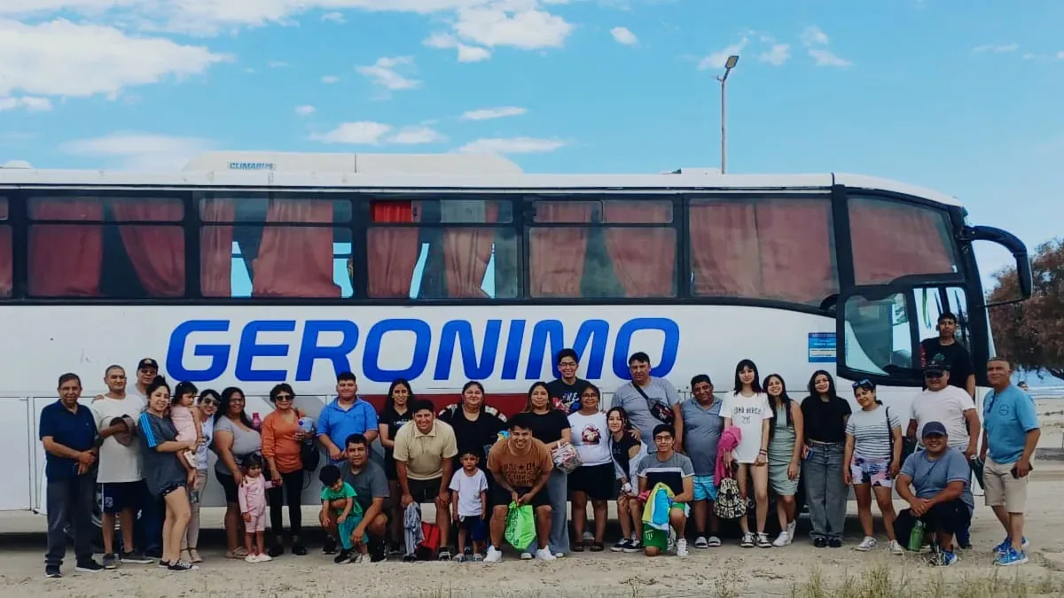 A large group of people poses happily in front of a white tour bus labeled "GERONIMO" under a clear blue sky.