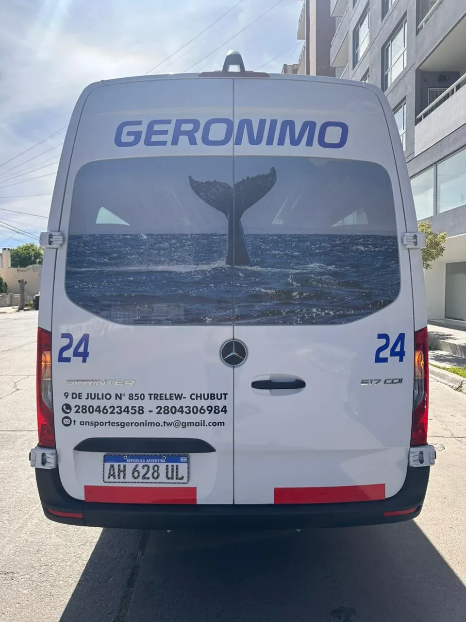 A white delivery van with bold, blue "GERONIMO" branding features a striking whale tail design amidst ocean waves on its rear, parked in an urban street by modern buildings.