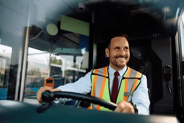 A smiling bus driver wearing a reflective safety vest sits behind the steering wheel in the cockpit of a city bus filled with natural light.
