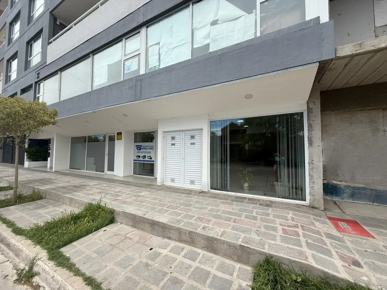 A modern, multi-story commercial building features large glass windows and a white facade, with a tree and neatly arranged paved walkway in the foreground.