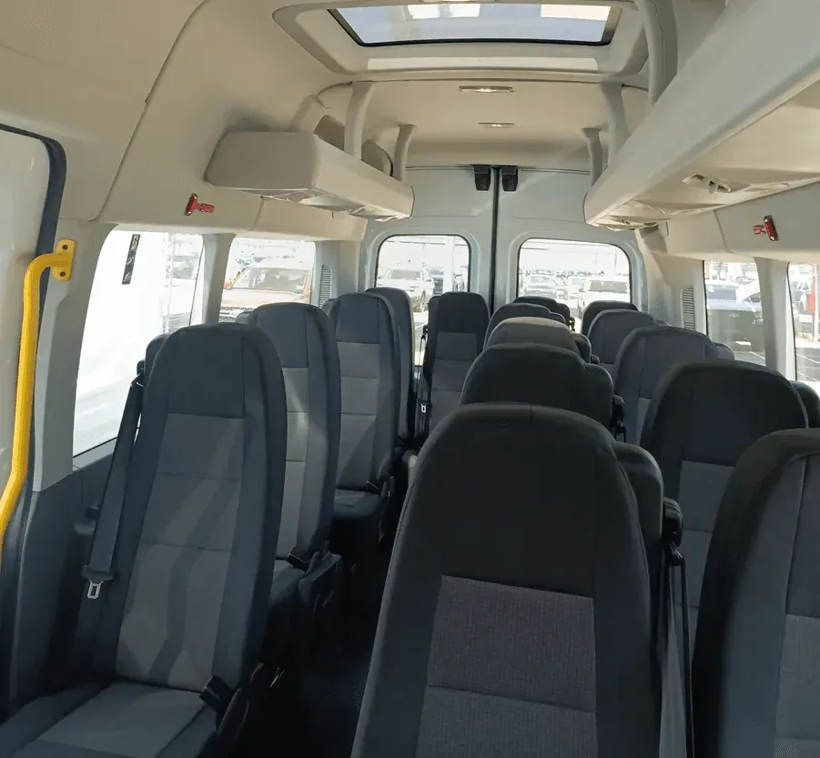 The interior of a spacious van features rows of comfortable, dark-gray seats with seatbelts, ample overhead storage, large windows for natural light, and a sunroof, ideal for group transportation or shuttle services.