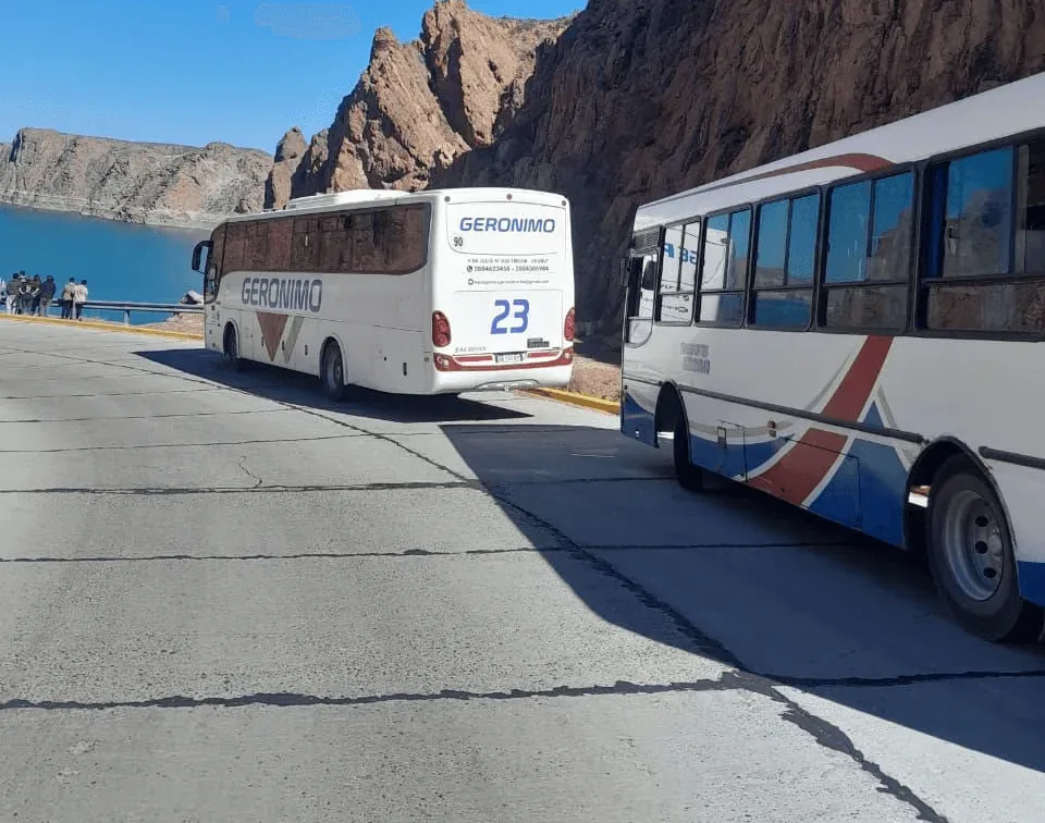 Two tour buses with red and blue stripes are parked on a mountain road alongside a serene blue lake, against a backdrop of rugged cliffs under a clear sky.